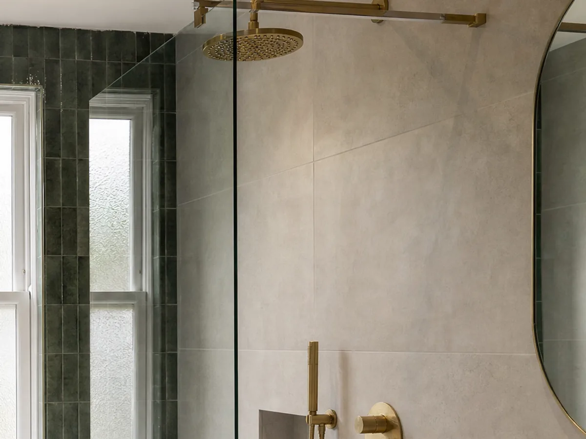 A walk-in shower with a clear glass panel, brass rainfall shower head and a recessed shelf